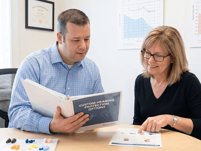 Audiology specialist reviewing a custom hearing protection options binder with a client, with sample earplugs and an audiogram visible nearby