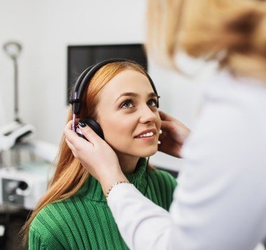 Patient wearing over-ear headphones during a speech audiometry test while a clinician assists with the fit
