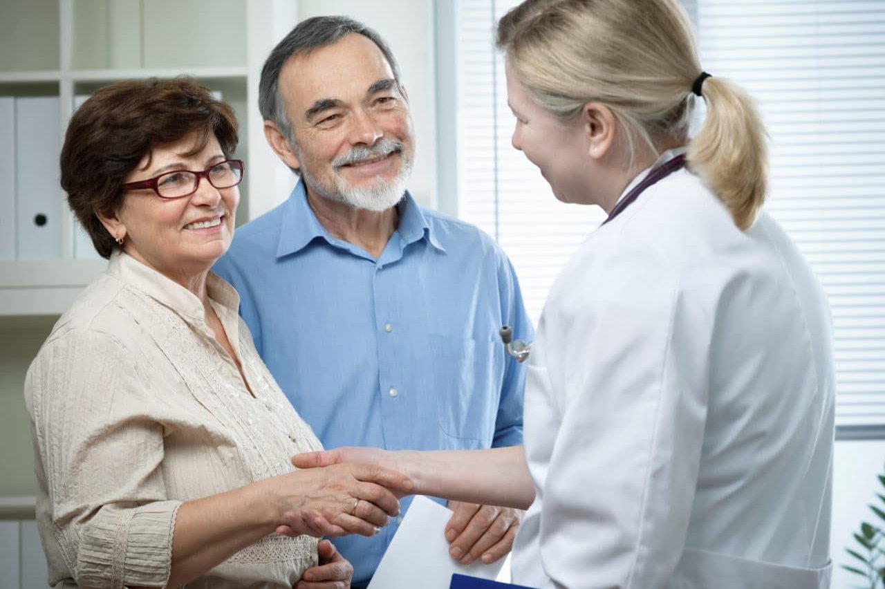 An audiologist and office team member greeting a smiling senior couple during a hearing consultation