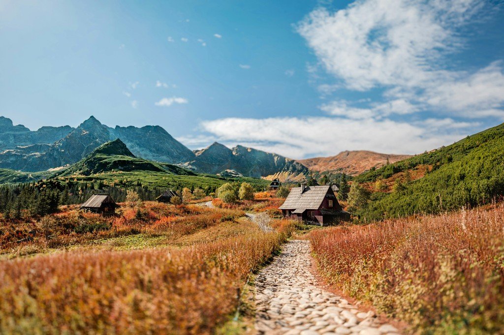 A stone pathway winds through a golden autumn mountain valley past small wooden cabins